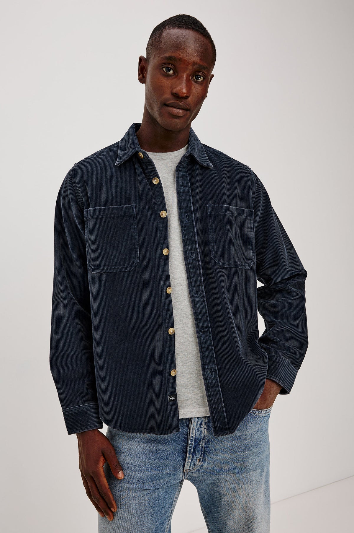 Man wearing a dark denim jacket over a gray shirt and light blue jeans on a white background