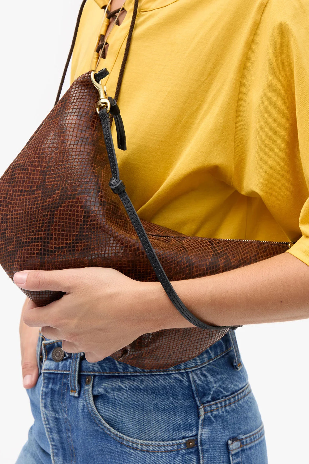 A person wearing a yellow top and blue jeans carries a brown snakeskin-patterned crescent shoulder bag with a black strap against a plain white background.