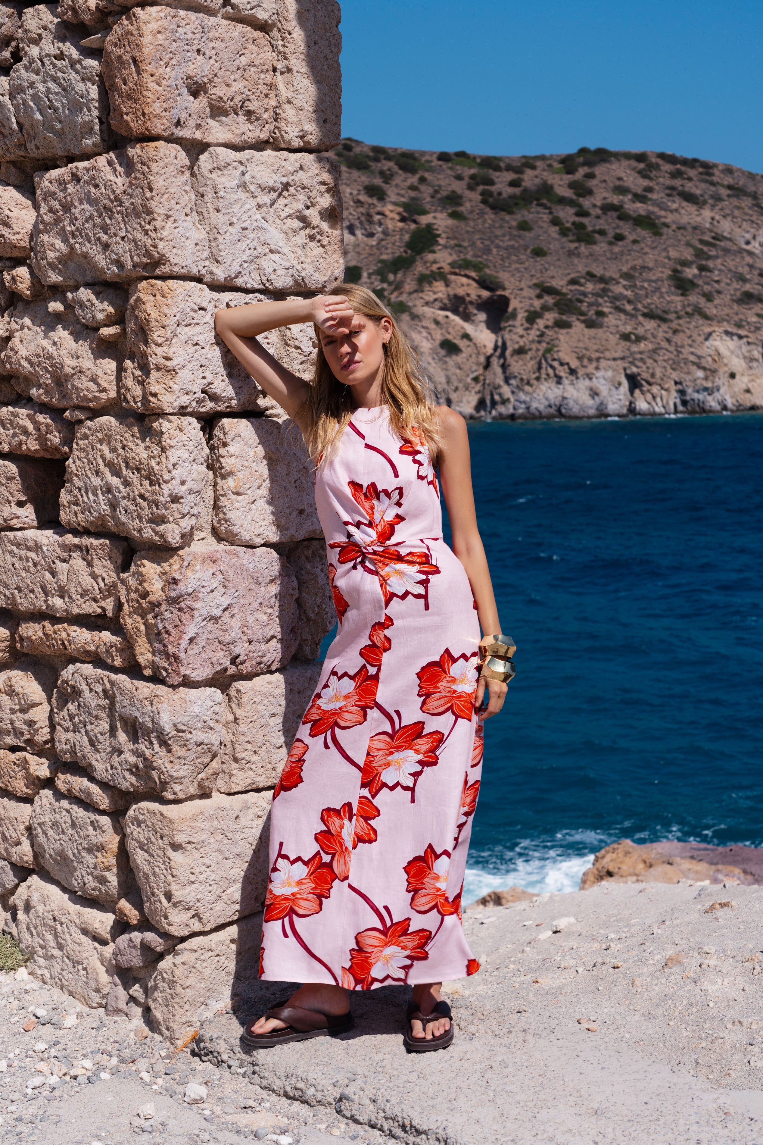 Woman in a floral dress standing against a stone wall with a scenic ocean view.