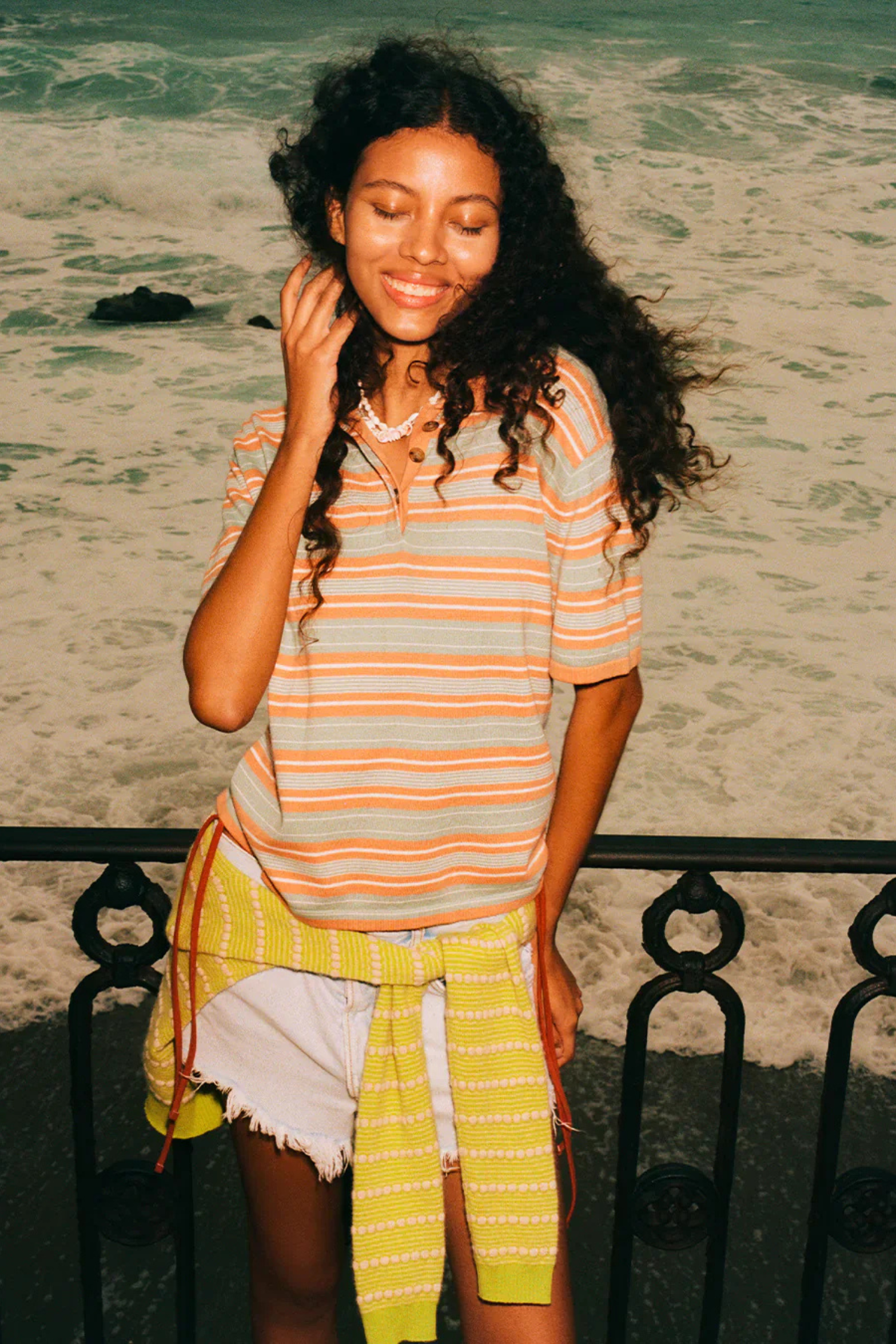 Woman in striped shirt and yellow skirt sitting on a railing by the beach.