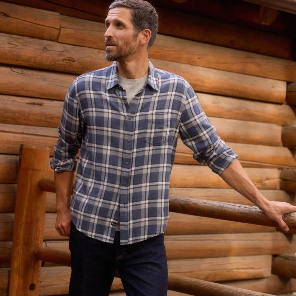 Man wearing a plaid shirt and dark pants standing against a wooden log cabin.