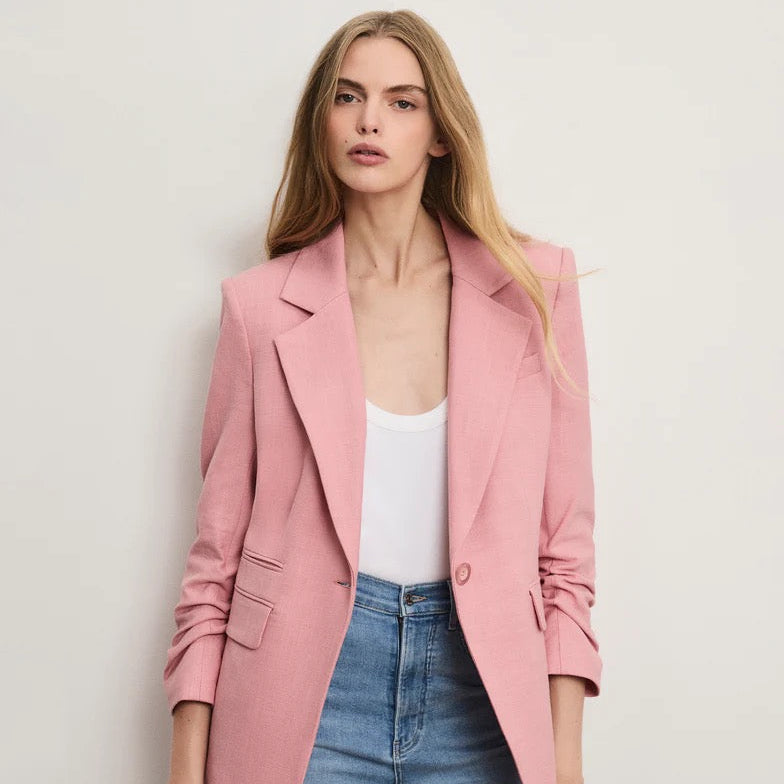 Woman wearing a pink blazer, white top, and blue jeans against a plain background