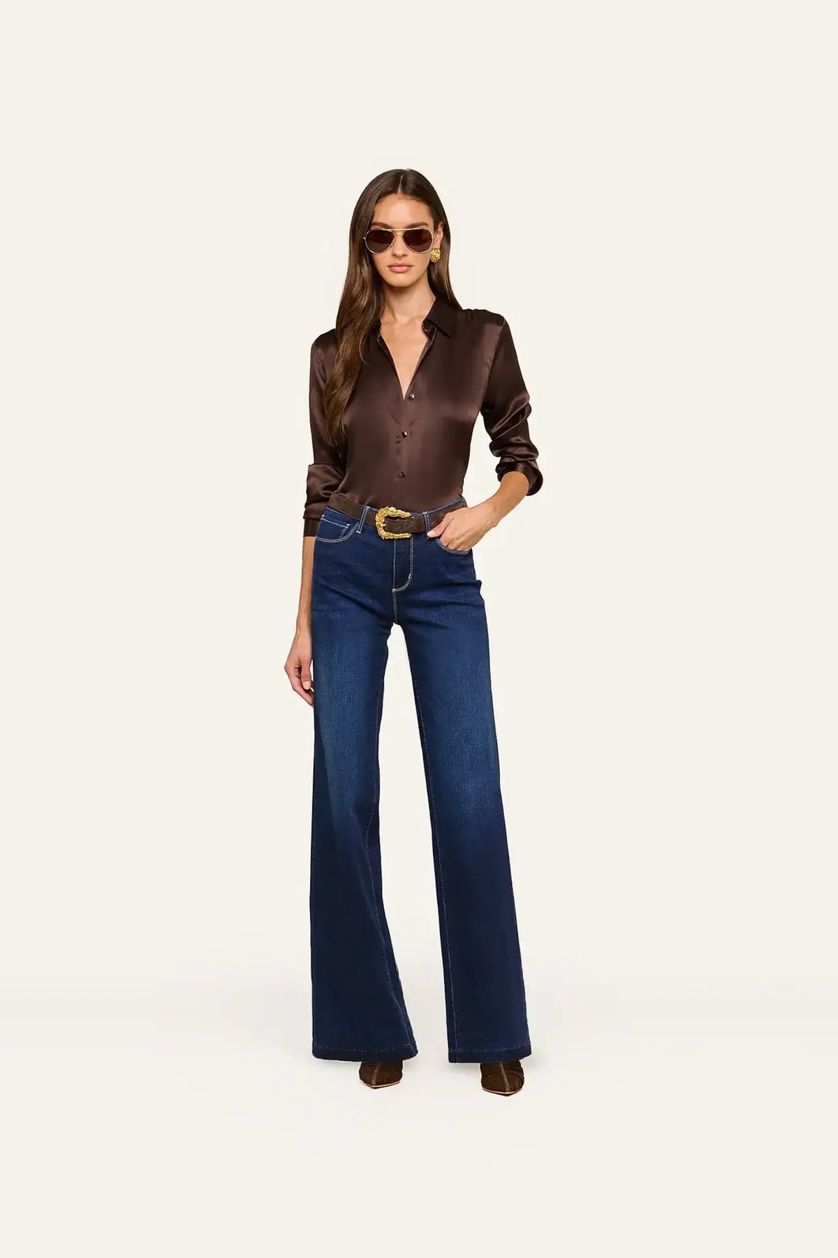 A woman modeling a dark brown satin button-up shirt and dark blue wide-leg jeans with a gold belt, standing against a plain off-white background.