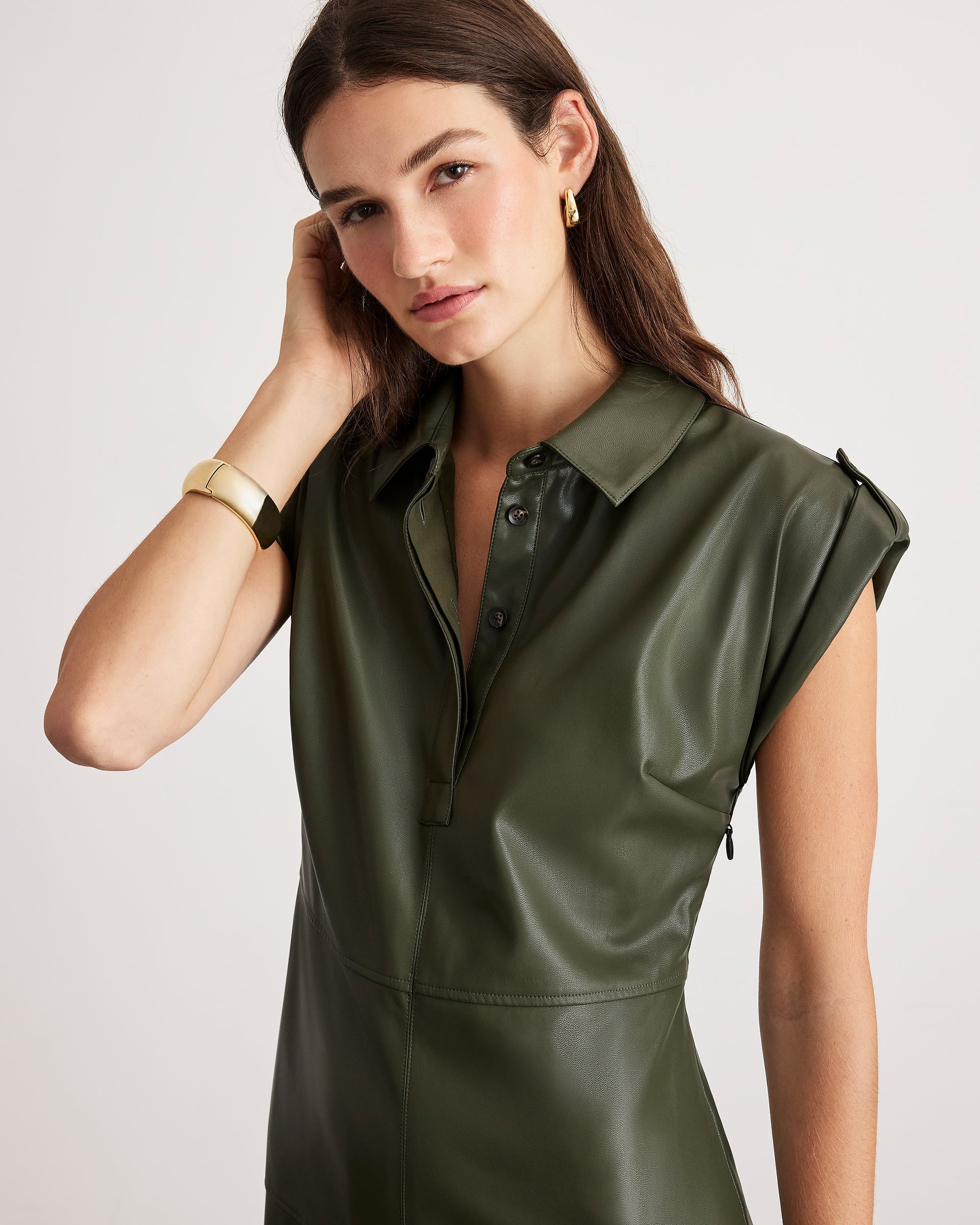 A woman wearing a dark olive green faux-leather collared shirt dress with cap sleeves and a buttoned placket, photographed against a light gray studio background.
