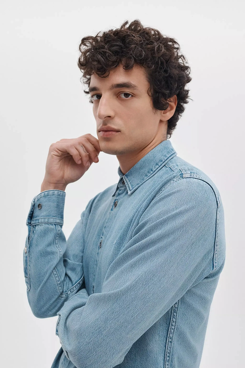 A man with curly brown hair wearing a light blue denim long-sleeve button-up shirt, posing against a plain white studio background.
