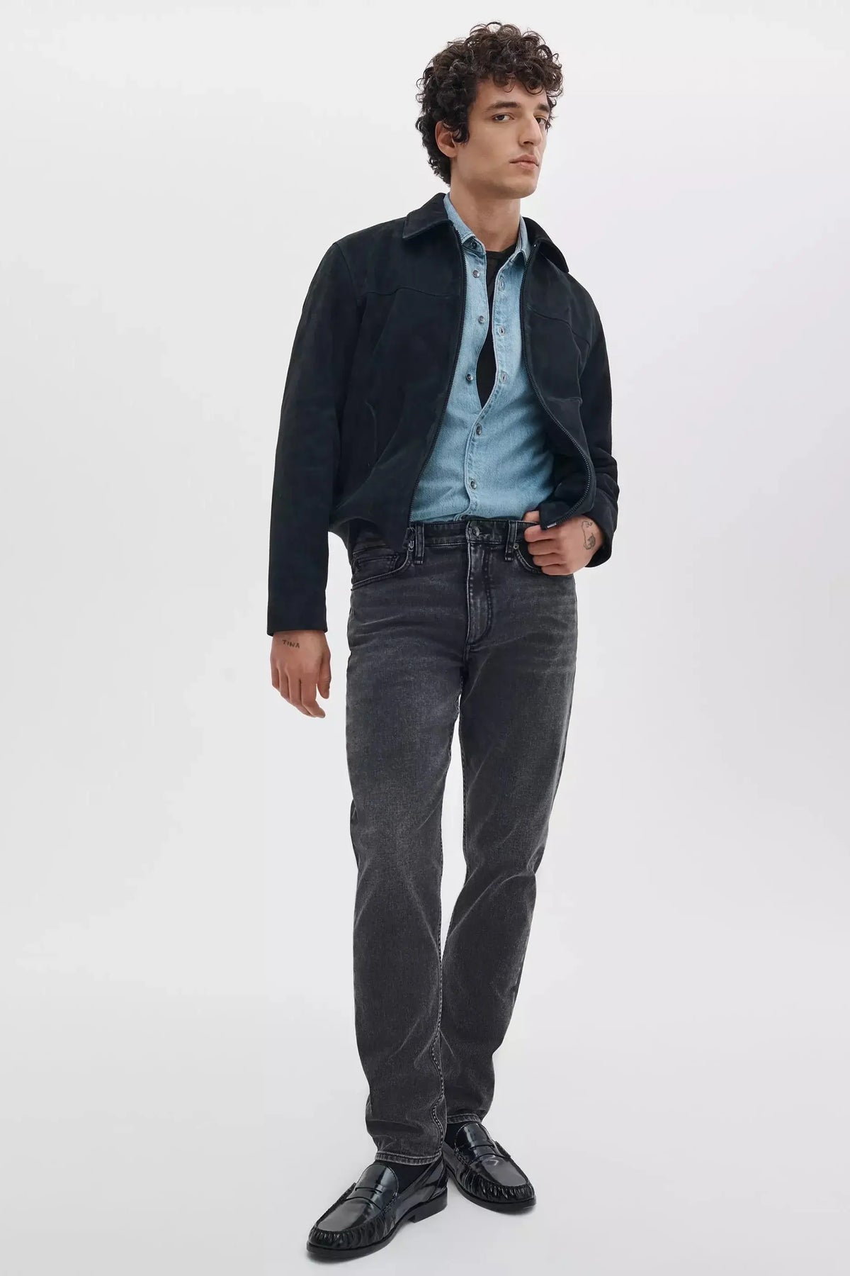 A man modeling a dark charcoal jacket over a light blue button-down shirt, paired with dark gray jeans and black leather loafers, against a plain white studio background.