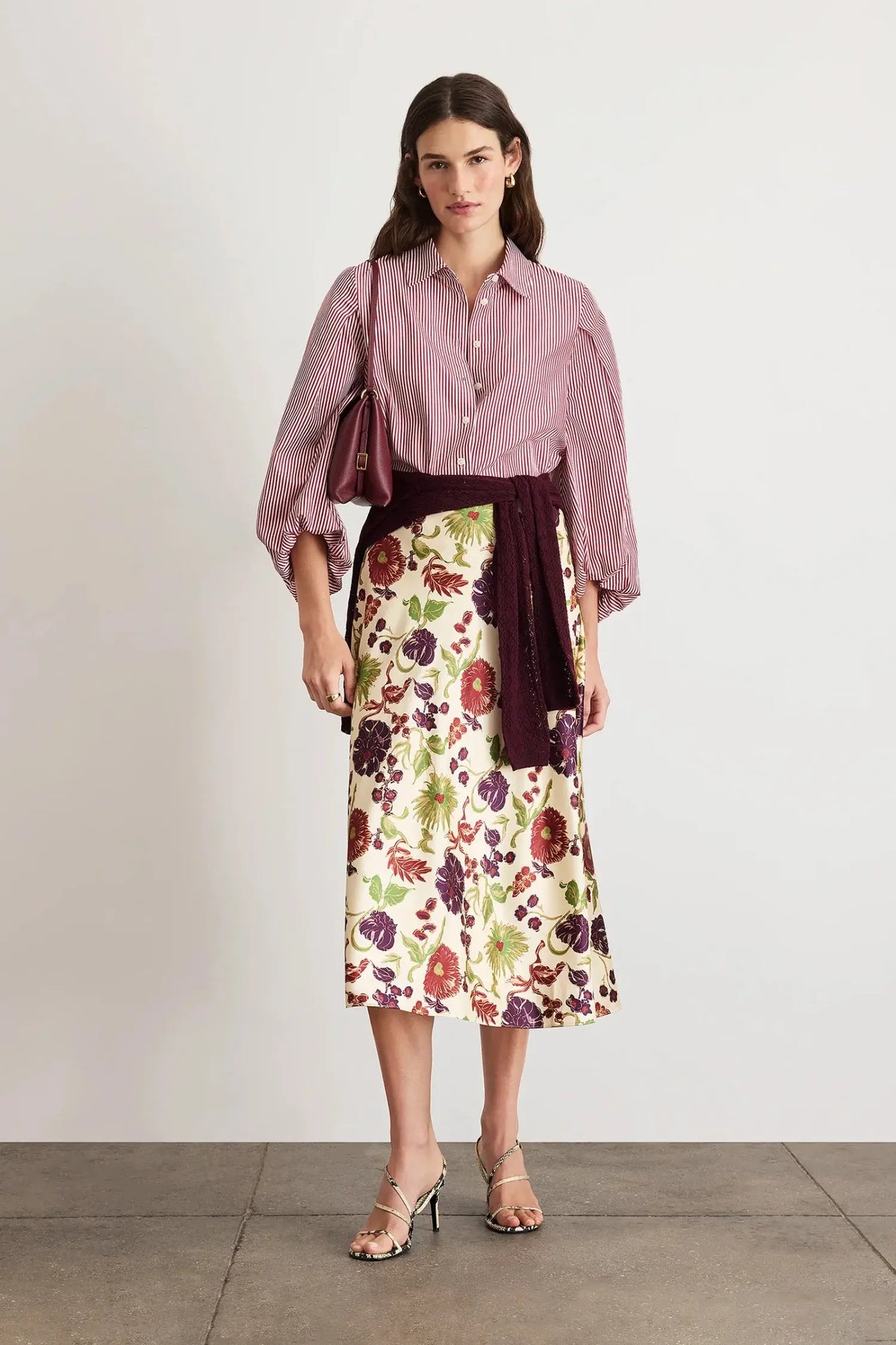 A woman stands against a plain light gray wall wearing a mauve button-up shirt with three-quarter sleeves, a cream midi skirt with a red and green floral print, and a dark burgundy velvet sash tied at her waist. She is also wearing clear high-heeled sandals and carrying a burgundy shoulder bag.